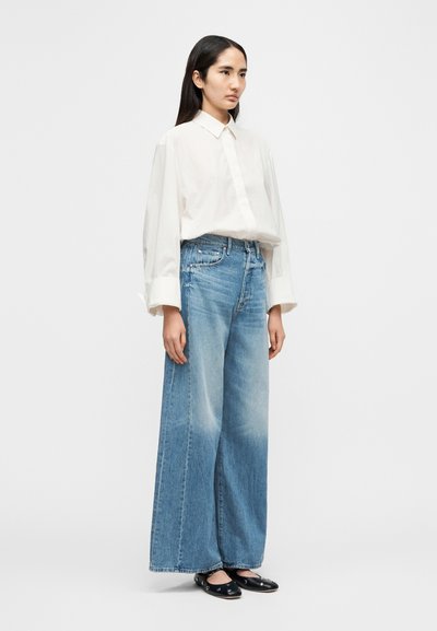 Λευκό πουκάμισο με μακριά μανίκια και κουμπιά στο εμπρός μέρος, συνδυασμένο με ανοιχτό μπλε τζιν παντελόνι φαρδιάς γραμμής και μαύρα παπούτσια slip-on. Απλός σχεδιασμός και εφαρμογή.