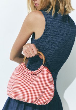 Donna in un abito blu navy senza maniche che tiene dietro la schiena una borsa a quadri rossa e bianca con manici tondi in legno.