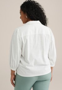 Camicia bianca, morbida e ampia, con maniche a sbuffo, colletto rotondo e dettagli di pieghe sul retro, abbinata a pantaloni verdi chiari. Tessuto liscio.
