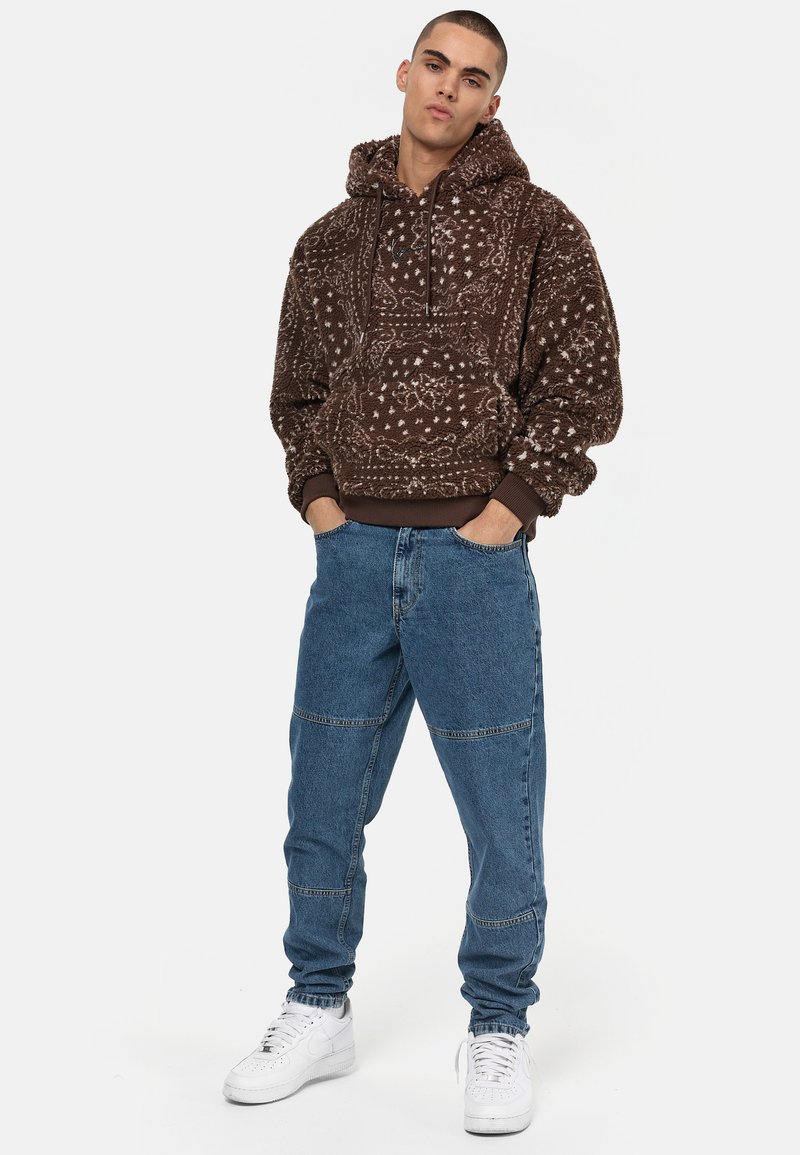 Brown hoodie with a textured pattern, featuring a drawstring hood and ribbed cuffs. Paired with light blue denim pants and white sneakers.