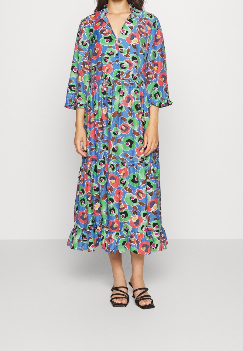 Femme portant une robe midi fleurie colorée avec un ourlet à volants et des manches trois-quarts, associée à des sandales noires à brides.