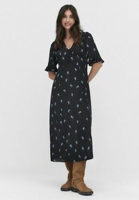 Schwarzes Blumenkleid mit V-Ausschnitt, kurzen Rüscheärmeln und verstreuten blauen Blumenmustern. Kombiniert mit braunen kniehohen Stiefeln.