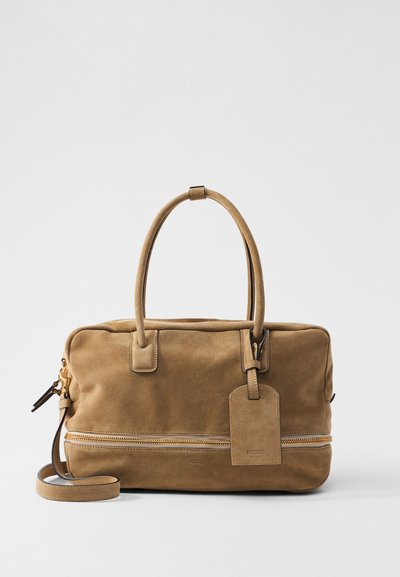 Geantă din suede bej cu mânere duale, buzunar cu fermoar și etichetă de bagaje. Textură netedă cu accente de hardware aurii și design minimalist.