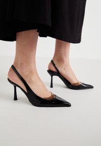 Décolleté slingback in vernice nera con punta affusolata e trasparenze sul davanti, posizionate su una superficie leggera e uniforme.