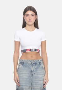 Maglietta bianca corta con maniche corte, caratterizzata da una fascia con logo "DIESEL" in nero e rosso in contrasto, posizionata all'orlo. Abbinata a pantaloni shorts in denim azzurro.