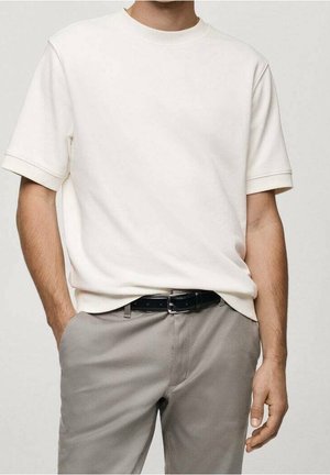 Homme portant une chemise à manches courtes écrue rentrée dans un pantalon gris clair avec une ceinture noire, debout avec une main dans la poche.