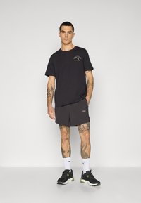 Sort bomulds T-shirt med et lille grafisk motiv, kombineret med mørke shorts. Modellen har sorte sportssko og hvide ankelstrømper på, der viser tatoveringer.