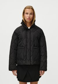 Black quilted jacket featuring a high collar, two front pockets, and a zip closure. Smooth texture with a puffy design and subtle pattern.