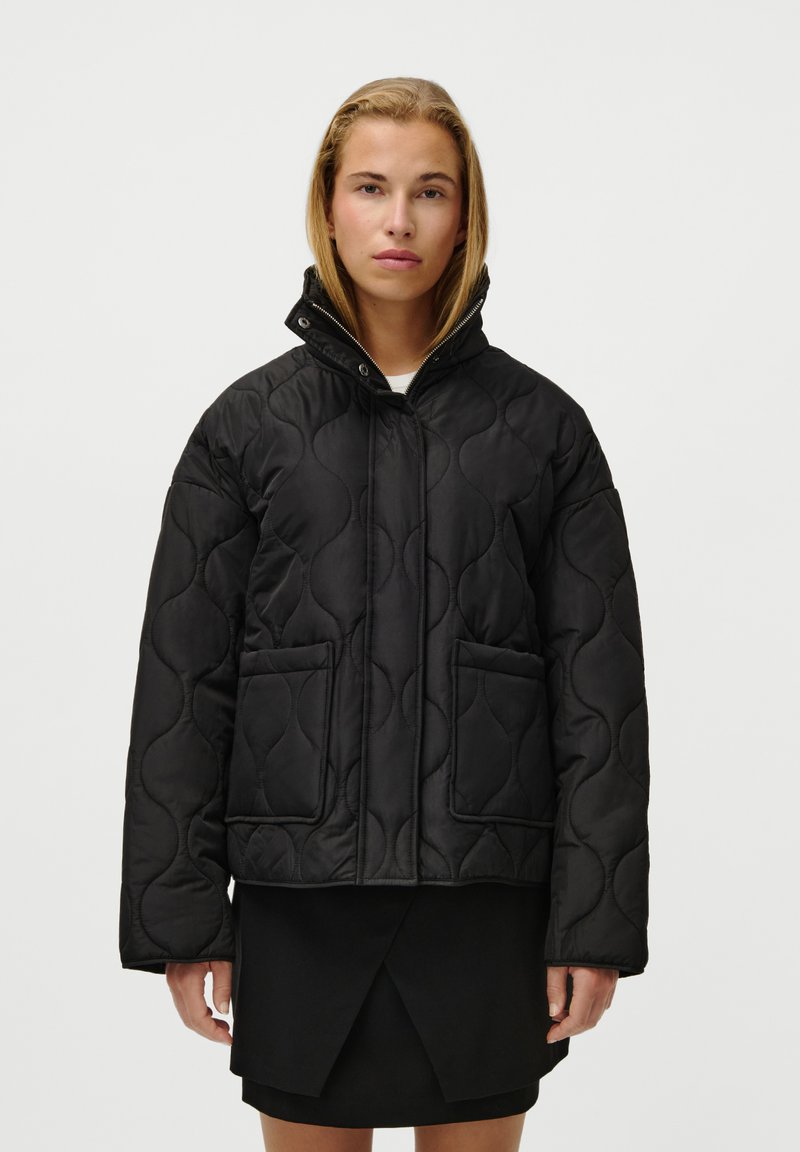 Black quilted jacket featuring a high collar, two front pockets, and a zip closure. Smooth texture with a puffy design and subtle pattern.