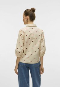Beige blus med röd hjärttryck, knäppbar krage, puffärmar och en avslappnad passform, ihop med lösa blå denimjeans.