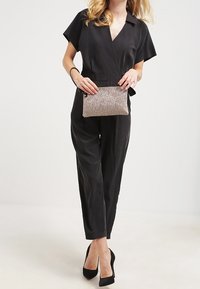 Salopette nera con maniche corte, colletto aperto e vestibilità rilassata. Modello tiene in mano una piccola borsa a tracolla metallica effetto texture con chiusura zip.