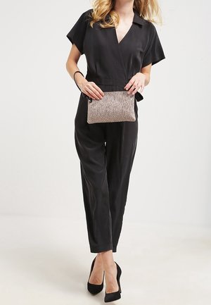 Combinaison noire à manches courtes, col ouvert et coupe décontractée. Le mannequin tient une petite pochette métallique texturée avec fermeture éclair.