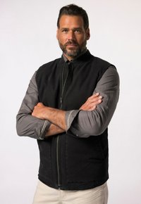 Chaleco negro con cremallera frontal y cuello, llevado sobre una camisa de manga larga gris. El chaleco tiene una textura suave, mientras que la camisa presenta puños decorativos.