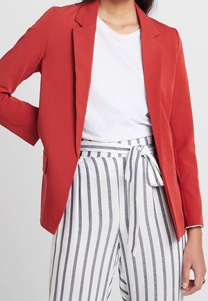 Femme portant un blazer rouge, un t-shirt blanc et un pantalon blanc taille haute avec des rayures verticales bleues nouées à la taille en un nœud.