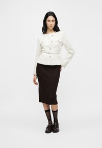 Veste en tweed blanche avec détails de boutons et poches avant, associée à une jupe midi côtelée marron, des socquettes noires transparentes et des mocassins foncés.