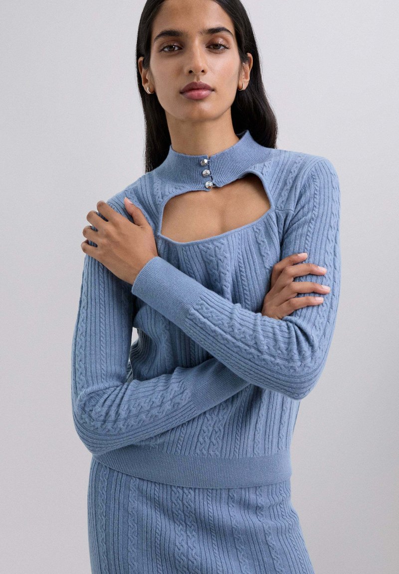 Suéter de punto azul claro con un cuello alto, con un corte rectangular y tres botones decorativos. Diseño de punto trenzado texturizado.
