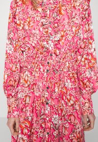 Robe fleurie rose vif avec devant à volants, boutons argentés au centre, taille élastique et manches longues bouffantes.