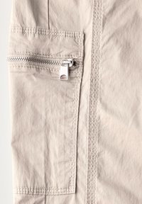 Beige stof met een gladde textuur heeft een zilveren ritsvak, gedetailleerde stiksels en een vierkante patch boven de rits.