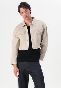 Beežist lühike denim jakk, millel on krae, kaks rinna taskut ja nupuga kinnitused. Kantud musta kudumtopi peal ja kombineeritud tumedate teksadega.
