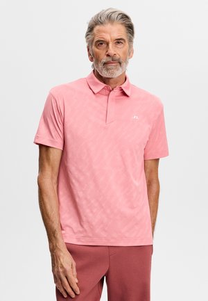 Älterer Mann mit grauem Haar und Bart, der ein lachsrosa kurzärmliges Polohemd und dazu passende Hose trägt, vor einem schlichten Hintergrund stehend.