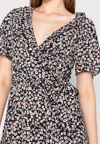 Femme portant une robe portefeuille noire avec un imprimé floral blanc, rouge et bleu, à manches courtes et à encolure en V froncée.