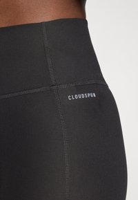 Crne tajice visokog struka izrađene od rastezljivog materijala s vidljivim šavovima i logotipom "CLOUDSPUN" sa strane. Glatka tekstura.