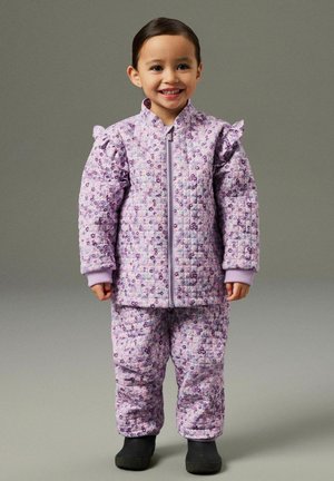 Copil zâmbitor purtând o geacă și pantaloni matlasați cu flori mov asortați, cu cizme negre, stând pe un fundal simplu.