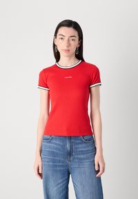 Raudona juostota t-shirt su juoda ir balta apdaila kaklo ir rankovėse. Ant krūtinės yra mažas logotipas. Sujungtas su mėlynomis džinsais.