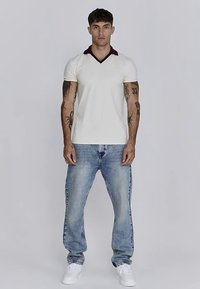 Vit kortärmad V-ringad tröja med vinröd krage, kombinerad med ljusblå jeans och vita sneakers. Modellen står vänd framåt.