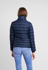 Granatowa kurtka puchowa z poziomym pikowaniem, wysokim kołnierzem i długimi rękawami, zestawiona z jasnymi dżinsami. Gładka faktura, casualowy design.