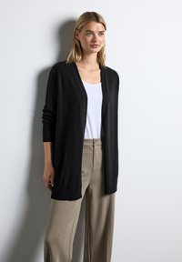 Czarny kardigan zapinany z przodu, wykonany z miękkiego materiału, z długimi rękawami i ściągaczami. Nałożony na biały top i beżowe spodnie.