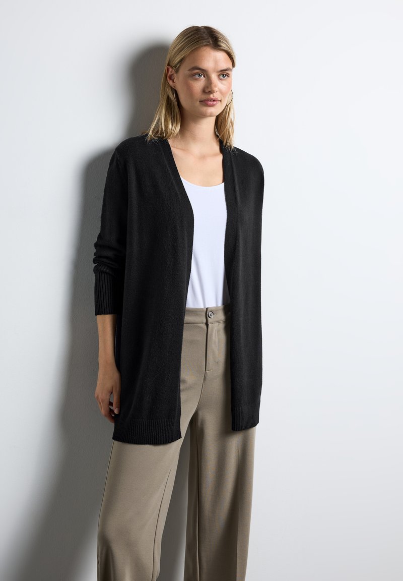 Czarny kardigan zapinany z przodu, wykonany z miękkiego materiału, z długimi rękawami i ściągaczami. Nałożony na biały top i beżowe spodnie.