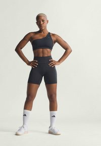 Top sportivo nero con design a spalla singola, abbinato a pantaloni corti neri a vita alta. Calzini bianchi con logo nero. Scarpe da ginnastica bianche.