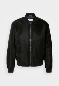 Schwarzige Bomberjacke aus glänzendem Nylon, mit einem Frontreißverschluss, gerippten Bündchen und Saum sowie einer Reißverschlusstasche am linken Ärmel.