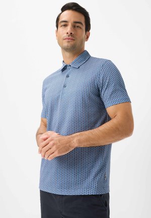 Mand iført blå mønstret kortærmet polo og mørke bukser, stående med hænderne foldet foran en hvid baggrund.