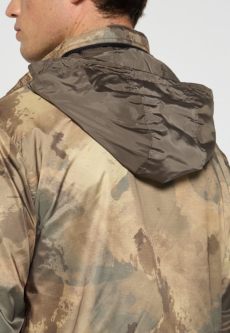 Gevoerde jas in bruin en beige camouflagepatroon; beschikt over een textuurstof en een ritskraag. Middengewicht ontwerp, geschikt voor gebruik buitenshuis.