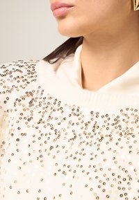 Cremefarbener, gestrickter Oberteil mit hohem Kragen, verziert mit goldenen und silbernen Pailletten, die ein strukturiertes Muster über den Stoff legen.