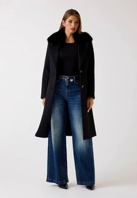 Długi czarny płaszcz z kołnierzem z futerka ekologicznego, zestawiony z szerokimi niebieskimi dżinsami. Płaszcz ma dopasowany krój i zapięcie na guziki.
