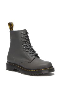 Bota de tobillo de cuero gris con seis ojales metálicos y cordones grises. Presenta costuras amarillas sobre una suela de goma negra y una tira de agarre en el talón.