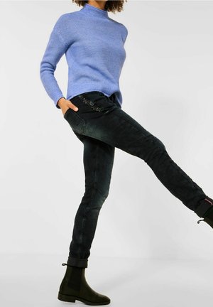 Personne portant un pull en maille bleu, un jean slim noir et des bottines noires, levant une jambe sur un fond blanc uni.
