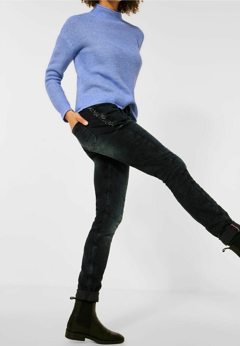 Personne portant un pull en maille bleu, un jean slim noir et des bottines noires, levant une jambe sur un fond blanc uni.