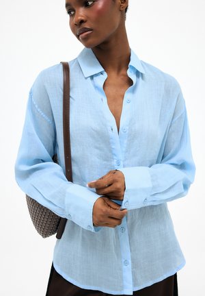 Žena v svetlomodrej košeli na gombíky s dlhými rukávmi, drží si manžetu a nesie hnedú tkanú tašku na ramene.