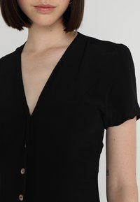 Černé šaty s výstřihem do V a krátkými rukávy, s přiléhavým střihem a knoflíky vpředu. Hladká textura látky.