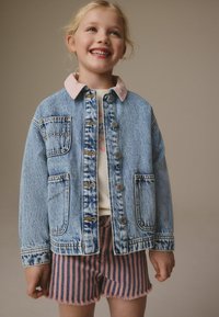 Chaqueta de mezclilla con lavado azul claro, cuello de pana rosa y cierre de botón. Combinada con pantalones cortos a rayas rosas y marinos con dobladillo deshilachado.