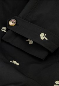 Zwarte stof met gouden borduursels met kleine bloempatronen. Een ronde knoop is bevestigd aan de manchette met een zichtbaar merklabel.