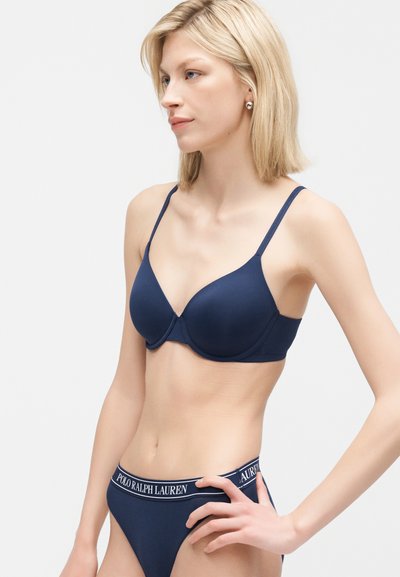 Γυναίκα με ξανθά μαλλιά που φοράει μπλε μποξεράκι και εσώρουχο με λωρίδα μέσης "Polo Ralph Lauren", στέκεται μπροστά σε απλό φόντο.