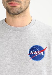 Grå sweatshirt med en rund blå NASA-logotyp som har vit text och en röd sväng, kort ribbad halsringning och en mjuk textur.