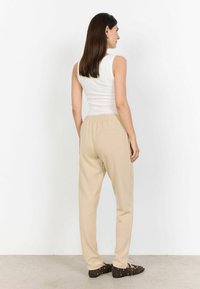 Beżowe spodnie dresowe z elastycznym paskiem w talii, bocznymi kieszeniami i gładką fakturą. Noszone z dopasowanym białym topem na ramiączkach i brązowymi butami.