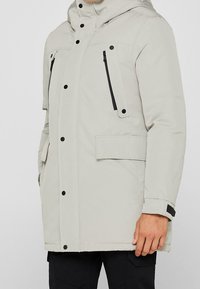 Giacca puffer grigia chiara con cappuccio, zip nere, tasche frontali e dettagli con bottoni a pressione. Texture liscia con isolamento imbottito.