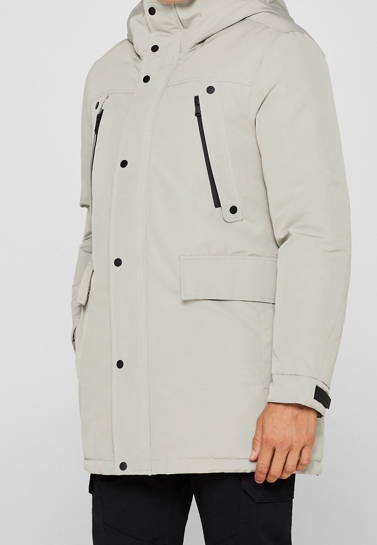 Giacca puffer grigia chiara con cappuccio, zip nere, tasche frontali e dettagli con bottoni a pressione. Texture liscia con isolamento imbottito.